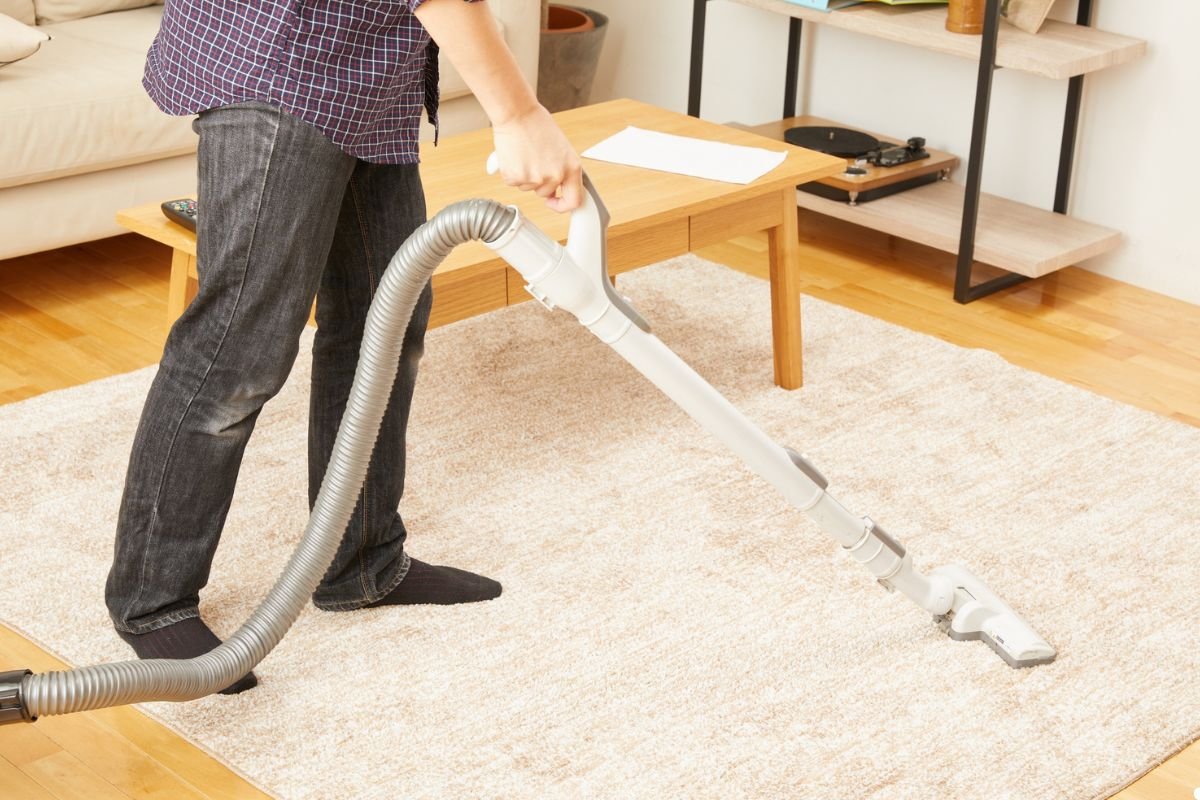 A man using a vacuum cleaner to clean a rug in a well-lit room.